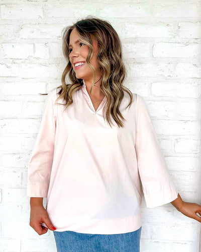 Woman wearing a light pink blouse and blue jeans against a white brick wall.