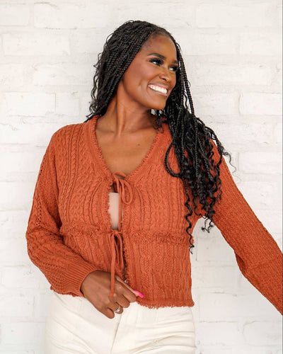Woman wearing a rust-colored knitted cardigan against a white brick wall