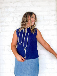 Woman wearing a blue sleeveless top with decorative buttons against a white brick wall.