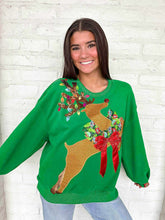 Load image into Gallery viewer, Woman wearing a green Christmas sweater with a reindeer design against a white brick wall.