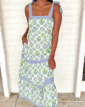 Load image into Gallery viewer, Woman wearing a floral dress with blue accents against a white brick wall.
